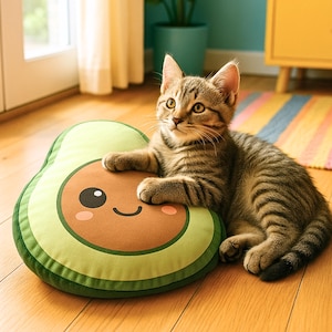 Puede incluir: Un gato atigrado descansa sobre un cojín verde con forma de aguacate y una cara sonriente. El gato tiene rayas marrones y negras. El cojín tiene un centro marrón y un borde verde.