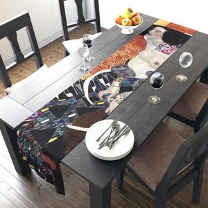 May include: A rectangular table runner featuring a print of a Gustav Klimt painting. The artwork depicts a nude figure with ornate patterns in shades of red, gold, and black. The runner is displayed on a dark wooden table, set with plates, silverware, and wine glasses.
