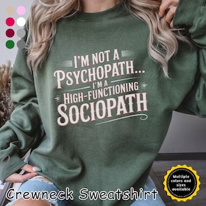 May include: A dark green crewneck sweatshirt with the text "I'M NOT A PSYCHOPATH... I'M A HIGH-FUNCTIONING SOCIOPATH" in white and red lettering. The sweatshirt has a round neckline and long sleeves. The image also shows a color palette and a badge that says "Multiple colors and sizes available."