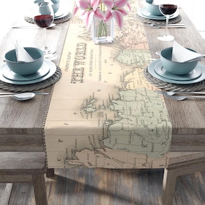 May include: A dining table set with a map-themed table runner. The runner displays a vintage world map in beige and green hues. The table is set with blue bowls, plates, and wine glasses. A vase of pink flowers provides a colourful contrast.