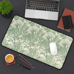 Puede incluir: Alfombrilla de escritorio rectangular con un estampado floral verde sobre un fondo crema. La alfombrilla tiene un borde negro y está rodeada por un portátil, un ratón, un cuaderno y una taza de café.