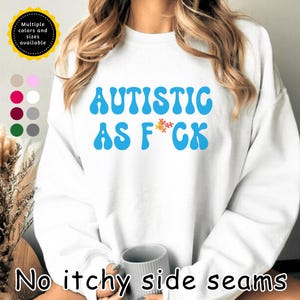 May include: White sweatshirt with the text "AUTISTIC AS F*CK" in blue, with a puzzle piece graphic. The sweatshirt has a crew neck and long sleeves. A color chart and text "Multiple colors and sizes available" are in the upper left corner.