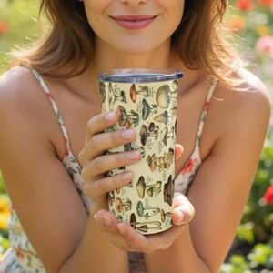 May include: A cylindrical travel mug with a cream-colored background and a pattern of various mushrooms in shades of brown, green, and orange. The mug has a dark blue lid and is held by a person.
