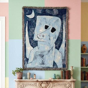 May include: A woven tapestry featuring an abstract figure in shades of blue and white against a dark blue background. The artwork includes a crescent moon and a textured border. The tapestry is displayed on a wall with a colorful, geometric design.