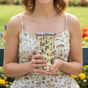 May include: A tall, insulated tumbler featuring a beige background with a detailed botanical illustration of various mushrooms in shades of brown, tan, and cream. The tumbler has a black lid and a stainless steel rim. The design is repeated on the matching dress.