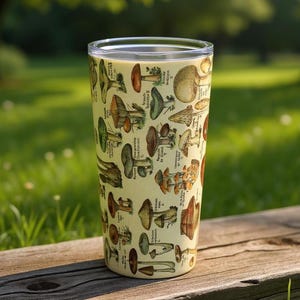 May include: A tall, clear tumbler with a cream-colored background and a repeating pattern of various mushroom illustrations in shades of brown, green, and orange. The tumbler is set on a wooden surface against a blurred green background.
