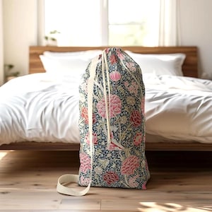 May include: A large laundry bag with a floral pattern in shades of navy, pink, and red. The bag has a drawstring closure and a cream-colored strap. The bag is standing on a wooden floor in front of a bed.