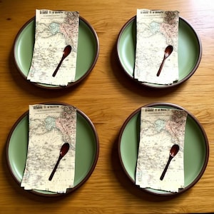 May include: Four green ceramic plates with brown rims, each holding a vintage map-printed napkin and a small wooden spoon. The plates are arranged on a light brown wooden surface, creating a coordinated table setting.