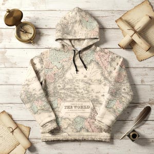 May include: A cream-coloured hoodie with a vintage world map design in pastel shades. The hoodie has a front pocket and a hood with a black drawstring. The text "THE WORLD" is visible on the front pocket. A compass, quill, and rolled-up papers are also present.