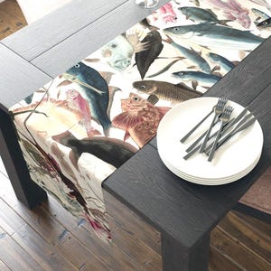 May include: A rectangular table runner featuring a watercolor illustration of various fish in shades of blue, pink, and brown on a white background. The runner is placed on a dark wooden table, with white plates and silverware.