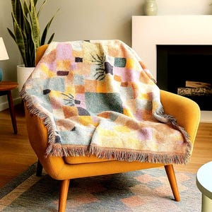 May include: A colorful woven throw blanket with a geometric pattern of squares and botanical designs in shades of orange, pink, green, and brown. The blanket is draped over a mustard yellow armchair, with a fringed edge.