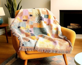 May include: A colorful woven throw blanket with a geometric pattern of squares and botanical designs in shades of orange, pink, green, and brown. The blanket is draped over a mustard yellow armchair, with a fringed edge.