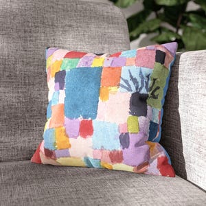 May include: Square throw pillow with a colorful abstract design. The pillow features a patchwork of watercolor-style blocks in shades of blue, red, yellow, green, and purple. The pillow is resting on a gray upholstered chair.