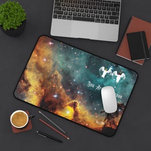 以下が含まれることがあります： 黄色、オレンジ、ティール色の鮮やかな星雲のデザインが施された長方形のマウスパッド。「You Are Gorgeous」の文字と2つの天使のグラフィックが描かれています。白いコンピューターマウスがパッドの上にあります。
