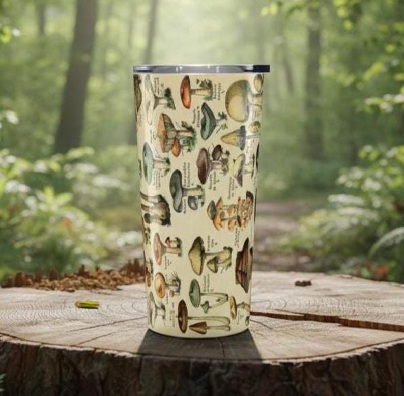 May include: A beige travel mug featuring a detailed illustration of various mushrooms in shades of brown, green, and yellow. The design includes scientific labels. The mug is cylindrical and has a silver rim and lid.