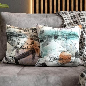 May include: Two decorative throw pillows with abstract art designs. One pillow features a design with black, purple, and tan elements. The other pillow has a design with blue, green, and orange shapes. Both pillows are on a gray sofa.
