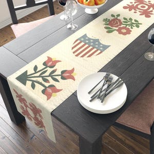 Könnte beinhalten: Ein cremefarbener Tischläufer mit einem sich wiederholenden Muster aus floralen und patriotischen Designs. Der Läufer zeigt rote, blaue und grüne Blumen- und Schildmotive. Der Läufer liegt auf einem dunklen Holztisch mit Tellern und Besteck.