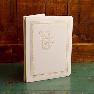 May include: White wedding guest book with a gold border and the text "Our Children's Wedding" in gold script.
