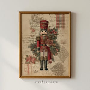Op de afbeelding: Ingelijste print met een vintage notenkrakersoldaat in een rood uniform, met een cadeau. De achtergrond bevat bladmuziek, poinsettia's en een ruitpatroon. De lijst is goudkleurig en de tekst "STUDIO PALETTE" staat onderaan.