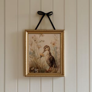 May include: A framed print featuring a duckling in a field of wildflowers. The artwork is in a gold-colored frame and is suspended by a black ribbon. The duckling is brown and white, and the flowers are yellow and white.