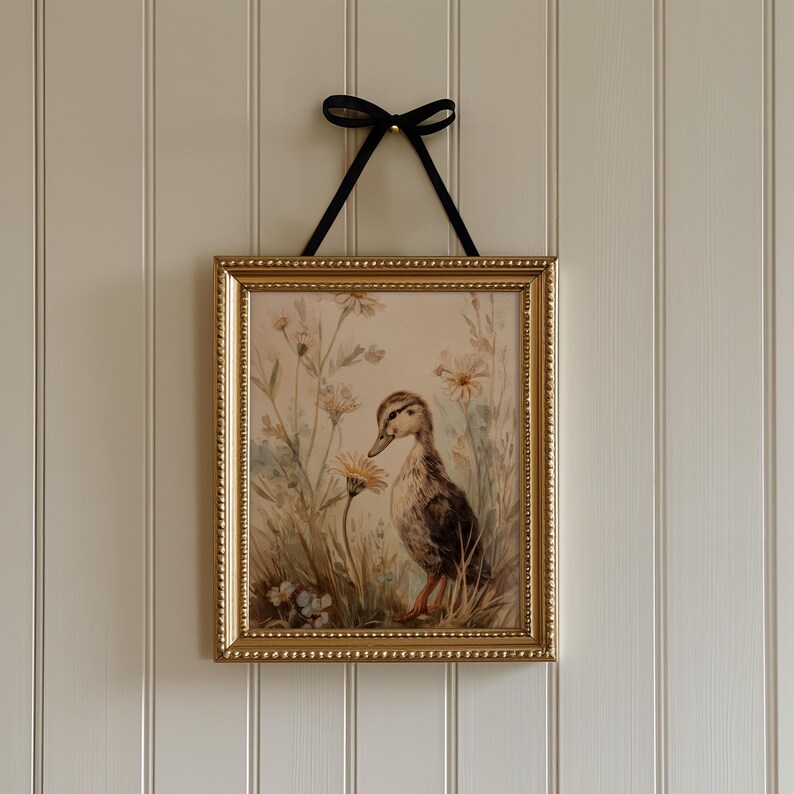 May include: A framed print featuring a duckling in a field of wildflowers. The artwork is in a gold-colored frame and is suspended by a black ribbon. The duckling is brown and white, and the flowers are yellow and white.