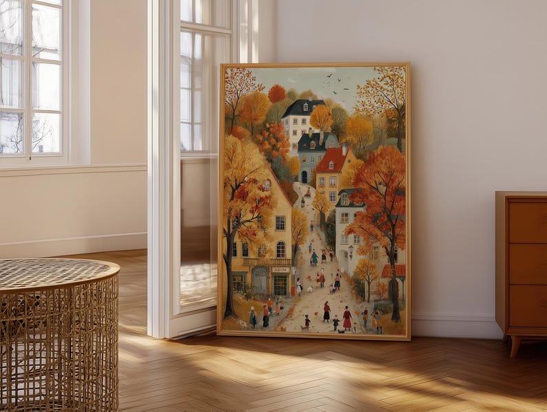 May include: A framed watercolor painting depicts a European village scene in autumn. The artwork features buildings with red and white roofs, surrounded by trees with orange and yellow foliage. People are walking on the street.