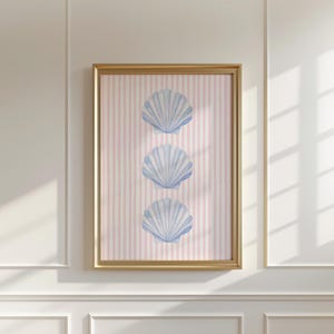 May include: A framed artwork featuring three blue seashell illustrations on a pink and white striped background. The artwork is encased in a gold-colored frame, creating a coastal or beach-themed aesthetic. The image is displayed on a white wall.
