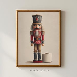 Könnte beinhalten: Gerahmter Druck mit einem klassischen Nussknacker in roter und goldener Uniform, neben einer Toilettenpapierrolle. Der Druck hat einen goldenen Rahmen und den Text "STUDIO PALETTE" unten.