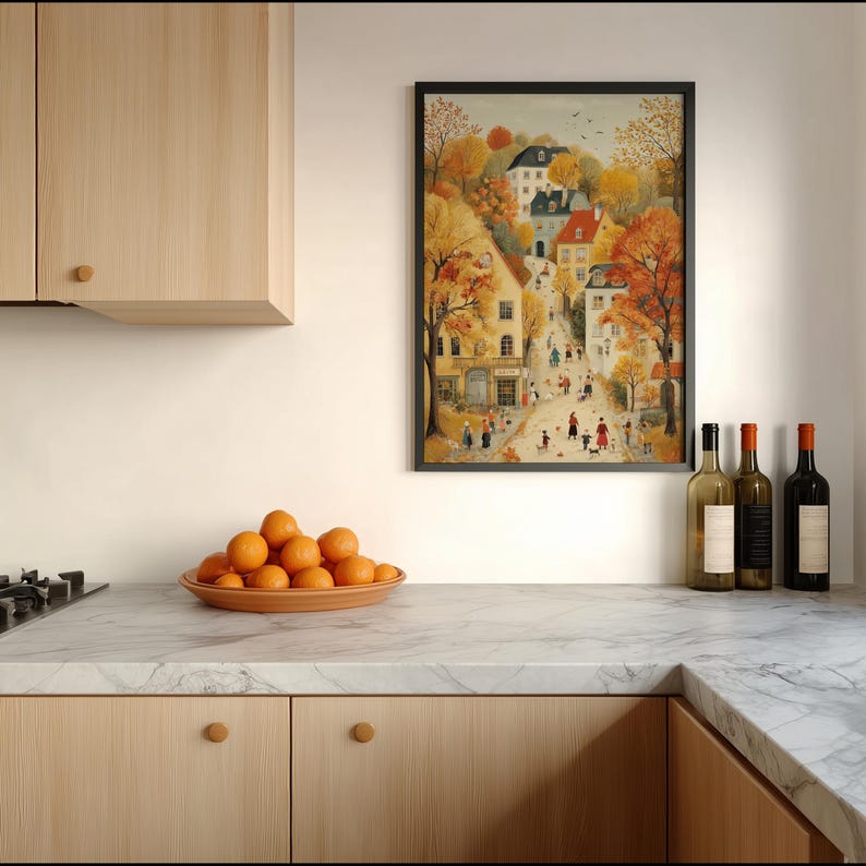 May include: A framed art print depicts a quaint European village scene in autumn colors. The artwork features buildings, trees with orange and yellow leaves, and figures walking along a street. Oranges are in a brown plate on a marble countertop.