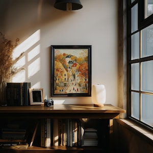 May include: A framed watercolor painting of a European village scene with autumn foliage. The artwork is displayed on a wooden shelf with books, a small framed photo, a camera, and a lamp. The room has a window with natural light.
