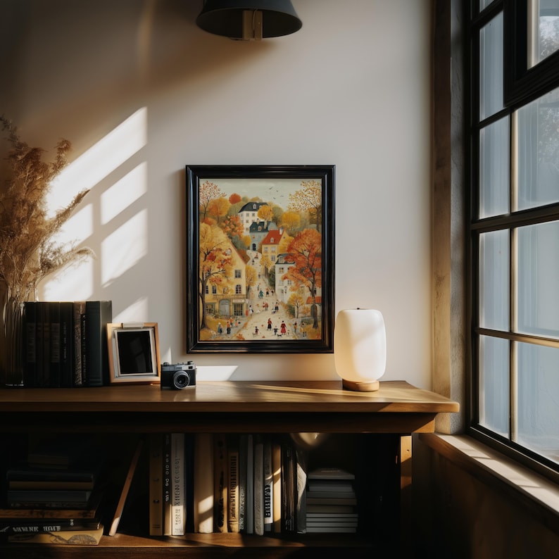 May include: A framed watercolor painting of a European village scene with autumn foliage. The artwork is displayed on a wooden shelf with books, a small framed photo, a camera, and a lamp. The room has a window with natural light.