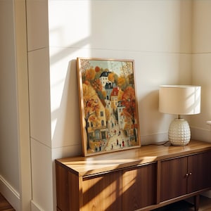 May include: A framed watercolor print of a European town in autumn. The artwork features colorful buildings, trees with orange and yellow leaves, and figures walking on the streets. The frame is a light brown color. The print is displayed on a wooden cabinet.
