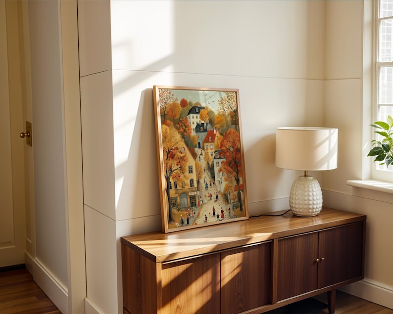 May include: A framed watercolor print of a European town in autumn. The artwork features colorful buildings, trees with orange and yellow leaves, and figures walking on the streets. The frame is a light brown color. The print is displayed on a wooden cabinet.