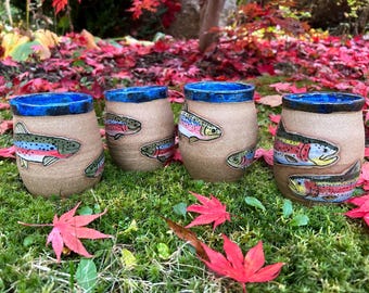 May include: Four ceramic mugs with a tan exterior and a vibrant blue interior rim. Each mug features a colorful fish design. The mugs are arranged on a bed of green moss and surrounded by red autumn leaves.