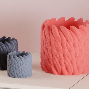 May include: Three decorative planters in shades of gray and coral. The planters have a textured, feather-like design around the exterior. The largest planter is coral, while the other two are gray.