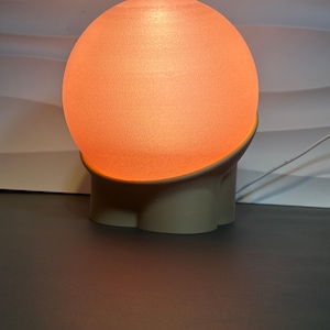 May include: A spherical lamp emitting a warm orange glow, resting in a matte beige base with a curved, molded design. The light source is a textured, translucent orb that sits securely within the solid, neutral-colored support structure.