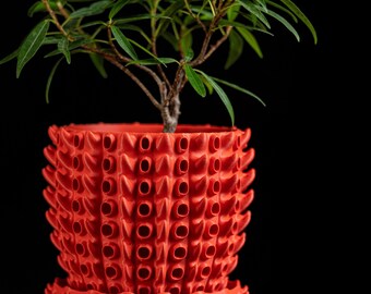 May include: A bright red, cylindrical plant pot with a repeating, textured, tubular pattern across its exterior surface. The pot includes a matching red saucer base and contains a small green plant with thin, pointed leaves.