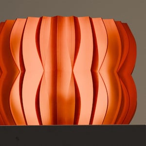 May include: A vibrant orange cylindrical vessel featuring a series of vertical, undulating, and deeply recessed pleats that create a rhythmic, wave-like pattern around the entire exterior surface of the object, resulting in a textured appearance.