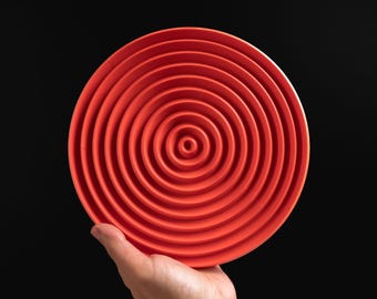 May include: A circular, bright orange silicone mat featuring a series of concentric, raised rings that radiate from a central point. The surface texture is uniform, creating a ripple-like pattern across the entire face of the round object.