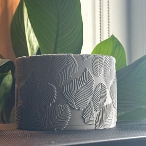 May include: A cylindrical gray planter featuring a raised, embossed pattern of multiple leaves covering the entire exterior surface. The texture consists of fine, parallel lines etched into each leaf shape, creating a tactile, repeating botanical design.