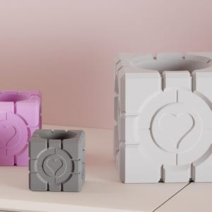 May include: Three cube-shaped planters in varying sizes and colors. The largest is white, the medium is lavender, and the smallest is gray. Each planter features a heart design in the center and a circular pattern around the heart.