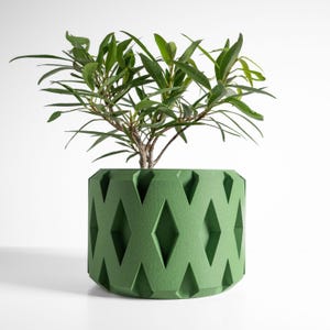 May include: A cylindrical green plant pot featuring a repeating geometric diamond pattern cut into the exterior surface. The pot has a matte, textured finish and contains a small green leafy plant with thin, pointed foliage.