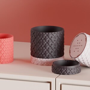 May include: Three cylindrical plant pots with a textured scale pattern on the exterior. One pot is coral, one is dark gray with a light pink base, and one is white, shown tilted to reveal a drainage hole pattern on the bottom base.