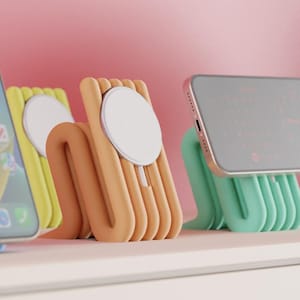 May include: Three U-shaped phone charging stands in pastel yellow, peach, and mint green. Each stand features a circular white magnetic charging disc integrated into the center, designed to hold a smartphone in a horizontal or vertical position.