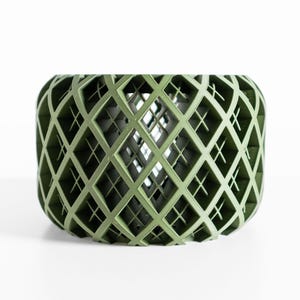 May include: A sage green, open-weave decorative bowl with a diamond lattice pattern. The bowl has a rounded shape and is set against a white background. The design allows light to pass through, creating shadows.