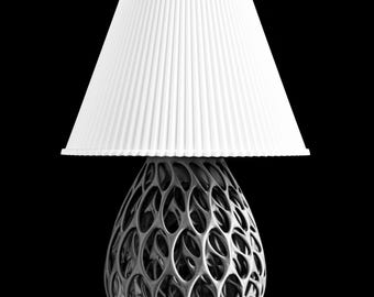 May include: A table lamp featuring a white, pleated, conical lampshade positioned atop a dark gray, egg-shaped base with an intricate, open-work lattice structure. The lamp rests on a square, dark, reflective base against a solid black background.