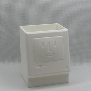 May include: A white 3D-printed desk organizer shaped like a vintage computer monitor. The front features a recessed screen area with a pixelated smiley face, a horizontal slot, and a small square icon in the bottom right corner.
