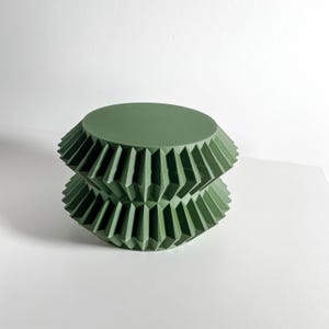 May include: A sage green cylindrical object featuring a flat circular top and a base with a repeating accordion-style pleated texture. The structure consists of two stacked, mirrored geometric sections with sharp, angular folds throughout the sides.