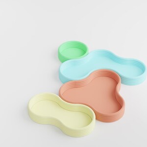 May include: A collection of four small, shallow trays with organic, amoeba-like shapes and ribbed side walls. The trays are colored in pastel shades of light green, sky blue, peach, and pale yellow, arranged in a cluster on a plain white surface.