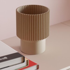 May include: A cylindrical beige vase with a vertically ribbed upper section and a smooth, solid base. The vase is positioned next to a stack of books, one of which features a graphic illustration of a red brain and the text Ben Lerner.